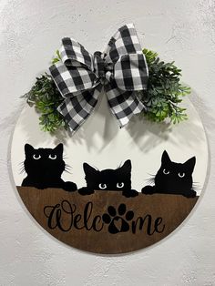 a welcome sign with three black cats and a checkered bow hangs on the wall