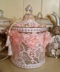 a pink lace covered container with flowers and pearls on it sitting on a table next to teapots