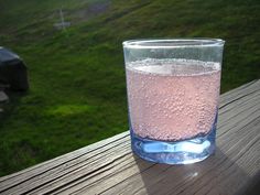 a glass filled with pink liquid sitting on top of a wooden table next to a green field