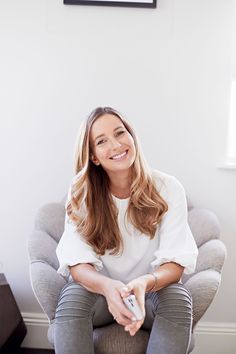 a smiling woman sitting in a chair with her legs crossed and holding a remote control