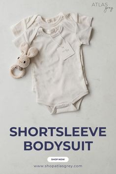 A flat-lay of two soft beige baby bodysuits with short sleeves, stacked neatly, accompanied by a crochet bunny rattle, all displayed on a light marble surface.