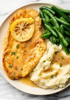 a white plate topped with chicken, mashed potatoes and green beans next to a lemon wedge