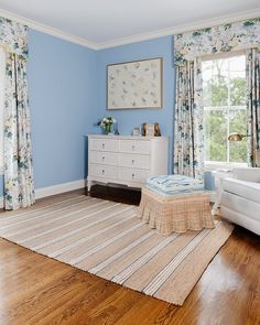 a bedroom with blue walls and floral curtains on the windowsills, a striped rug, white furniture, and wooden flooring