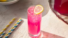 a pink drink in a tall glass with a slice of lemon on the rim and two straws next to it