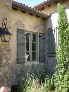 an old stone house with shutters open on the outside and green plants in front