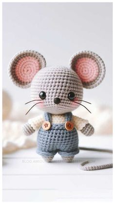 a small crocheted mouse is standing on a white surface and wearing overalls