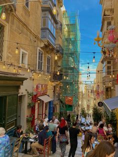 people are sitting at tables in an alleyway with many lights strung from the buildings