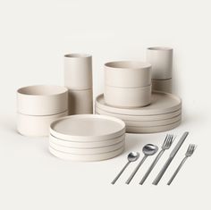 a set of white dishes and silverware on a white surface with spoons, forks and knives