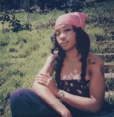 a woman sitting in the grass wearing a pink bandana and looking at the camera