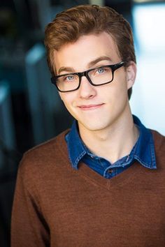 a young man wearing glasses and a brown sweater looks at the camera with an intense look on his face