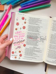 a person holding an open bible next to colored markers and pencils on a table