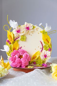 a white cake with pink and yellow flowers on it sitting on a table next to plates