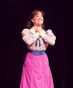 a woman in a pink dress standing on stage with her arms folded and smiling at the camera