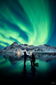 two people standing in front of an ice covered lake under the northern sky with green and blue lights