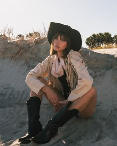 a woman sitting in the sand wearing a cowboy hat and boots with her legs crossed