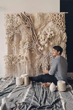 a man sitting on top of a bed in front of a wall hanging made out of yarn