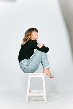 a woman sitting on top of a white stool in front of a gray wall smiling