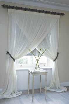 a white table sitting under a window next to a sheer curtain with flowers in it