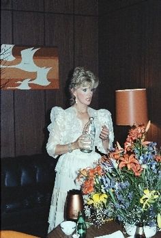 a woman standing in front of a table with flowers on it and a lamp next to her