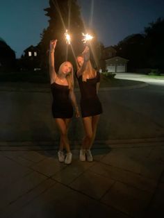 two young women holding sparklers in their hands on the sidewalk at night, both wearing short black dresses