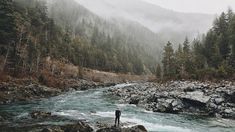 a man standing on rocks in the middle of a river surrounded by trees and mountains