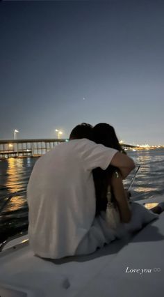 two people sitting on the back of a boat at night looking out over water with a bridge in the background