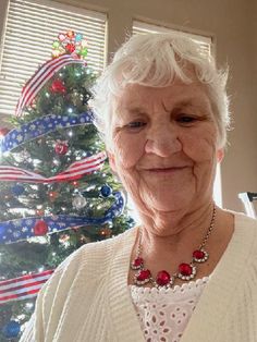 an older woman standing in front of a christmas tree wearing a white sweater and red beaded necklace