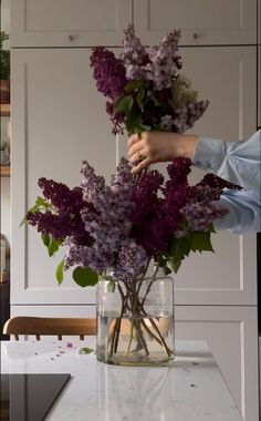 a vase with purple flowers in it sitting on a kitchen counter next to a person's hand