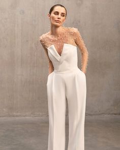 a woman standing in front of a concrete wall wearing white pants and a sheer top