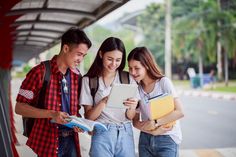 three college students looking at something on a tablet while standing in front of a bus stop