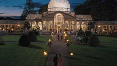 people are walking around in front of a glass house at night with candles on the walkway