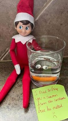 an elf sitting next to a glass filled with liquid and a note on the floor