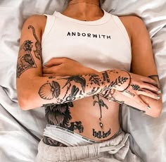 a woman laying in bed with tattoos on her stomach and arms, wearing a white tank top