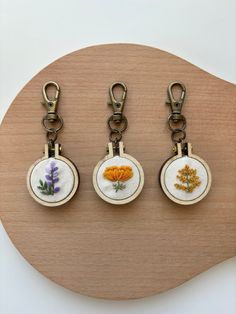 three key chains with embroidered flowers on them sitting on top of a wooden board in front of a white wall