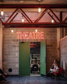 two people sitting in chairs under a neon sign that reads theatre on the wall behind them