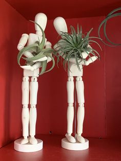 two white wooden figures holding plants in their hands on a red shelf against a red wall