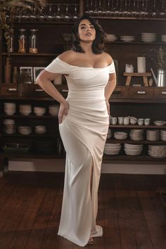 a woman wearing a white dress standing in front of a shelf with dishes on it