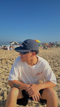 a man sitting in the sand on top of a beach wearing a hat and shorts