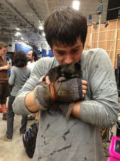 a man holding a small kitten inside of a building with other people in the background