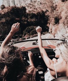 two women sitting in a convertible car with their hands up to the sky and one woman standing behind them