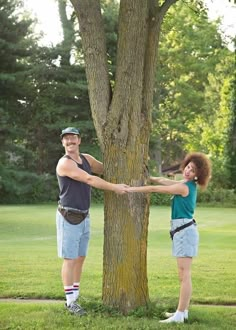 a man and woman standing next to each other near a tree with their hands on the trunk