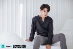 a young man sitting on top of a white bed next to a wall with vertical blinds