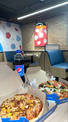 two pizzas in boxes sitting on a table next to a bottle of pepsi soda