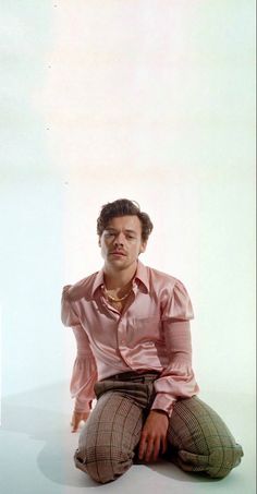 a man sitting on the ground in front of a white wall wearing a pink shirt and plaid pants