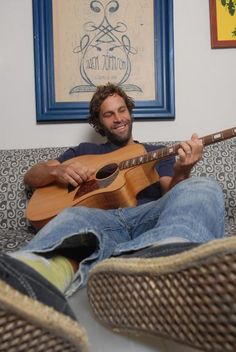 a man sitting on top of a couch with a guitar in his hand and smiling at the camera