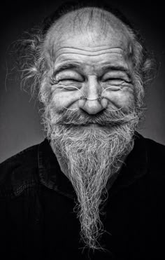 an old man with a long beard and big eyes smiles into the camera while wearing a black shirt