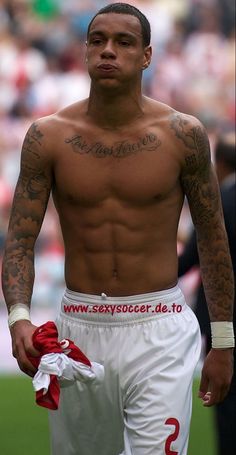 a shirtless man with tattoos on his chest and hands in his pockets, walking across a soccer field