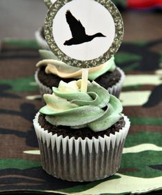 two cupcakes with green frosting and a black bird on top are sitting on a camouflage tablecloth