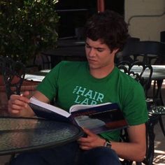 a young man sitting at an outdoor table reading a book