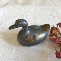 a metal duck sitting on top of a bed next to dried flowers and a white tablecloth